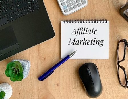 Affiliate marketing words on a notebook. With pen, mouse, calculator, plant, laptop and brown table background.